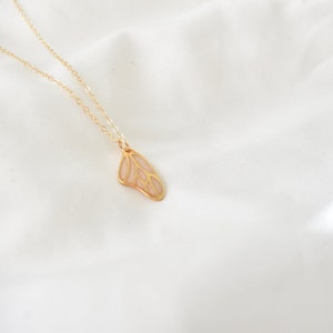 Puede incluir: Un delicado collar dorado con un colgante en forma de ala de mariposa. El ala tiene un relleno de esmalte rosa p&aacute;lido y est&aacute; suspendida de una fina cadena. El collar se muestra sobre un fondo blanco texturizado.