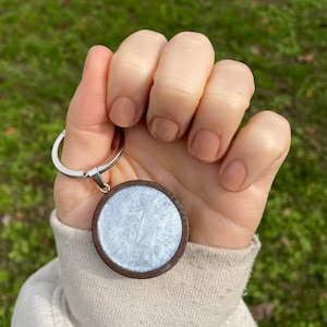Peut inclure: Un porte-clés rond en bois avec une surface blanche et chatoyante. Le porte-clés a un anneau en métal argenté.
