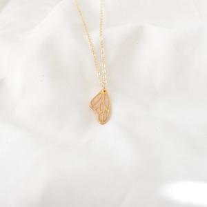 Puede incluir: Un delicado collar dorado con un colgante en forma de ala de mariposa. El ala tiene un relleno rosa claro con un patr&oacute;n de venas detallado. El collar se muestra sobre un fondo blanco texturizado.