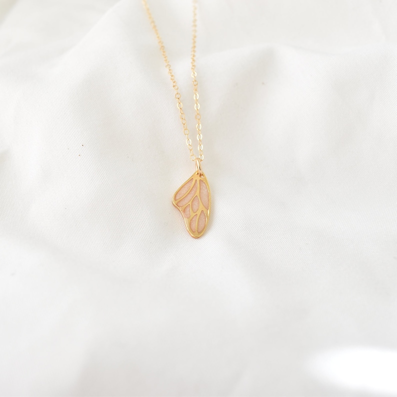 Puede incluir: Un delicado collar dorado con un colgante en forma de ala de mariposa. El ala tiene un relleno rosa claro con un patr&oacute;n de venas detallado. El collar se muestra sobre un fondo blanco texturizado.
