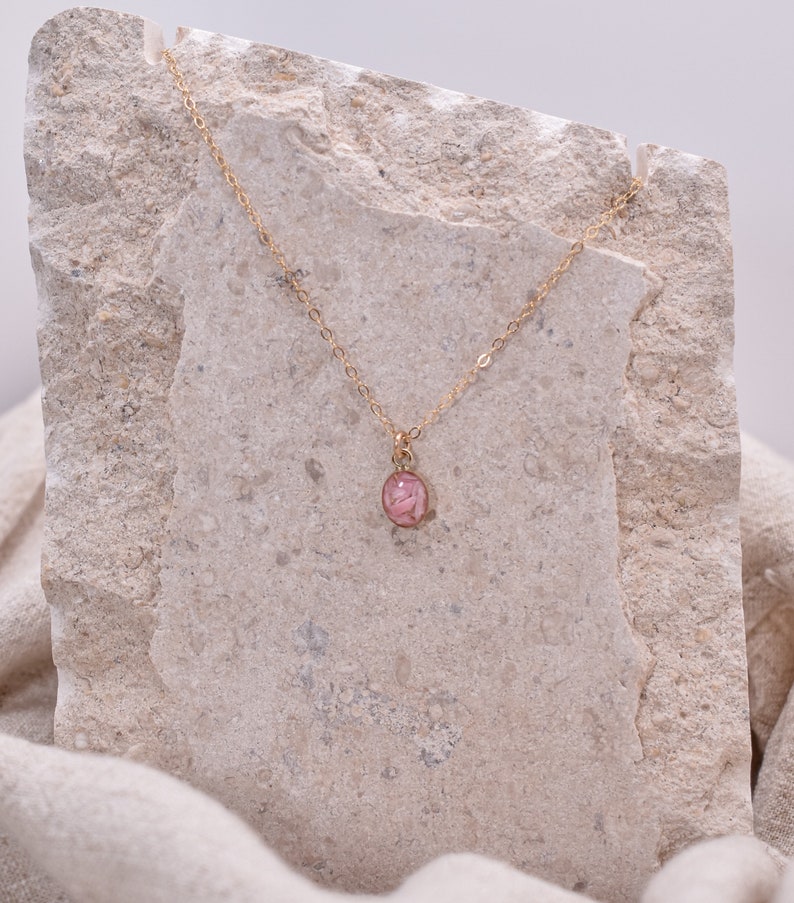 Op de afbeelding: Een delicate gouden ketting met een klein, rond hangertje met een geperste roze bloem ingekapseld in helder hars.