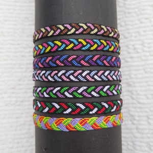 Op de afbeelding: Een set van acht kleurrijke vriendschapsarmbanden met een gevlochten patroon. De armbanden zijn gemaakt van draden in verschillende kleuren en zijn op elkaar gestapeld.