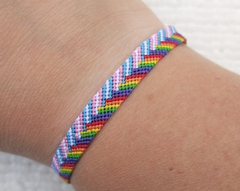 Puede incluir: Una colorida pulsera de la amistad con un patrón de chevrón. La pulsera presenta un arcoíris de colores, incluyendo rojo, naranja, amarillo, verde, azul y morado, así como rosa y blanco. La pulsera está hecha de hilos tejidos.