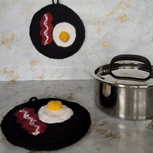 May include: Two black crocheted pot holders shaped like a fried egg and bacon. The pot holders are on a white countertop with a stainless steel pot in the background.