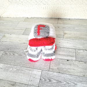 May include: A handmade crochet baby set in white, red, and gray. The set includes a hat with a gray square and a red letter "F", and matching booties. The booties and hat have red accents.