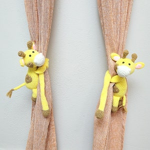 May include: Two crocheted yellow giraffe toys hanging from brown curtains. The giraffes have black eyes and brown spots.