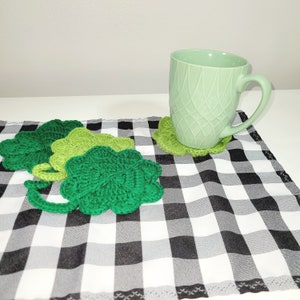 May include: Four green crocheted shamrock-shaped coasters on a black and white checkered tablecloth with a light green mug on top of one of the coasters.