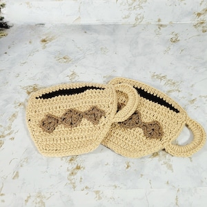 May include: Two crocheted coffee cup-shaped baskets with brown accents. The baskets are made with beige yarn and have a brown crocheted design on the front.