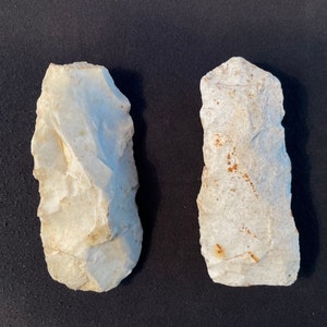 Puede incluir: Dos herramientas de piedra blanquecina en forma de mano, con bordes ásperos y astillados. Las herramientas se colocan una al lado de la otra sobre un fondo oscuro. Las herramientas de piedra parecen ser artefactos antiguos.