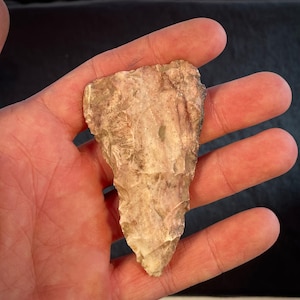 Pode incluir: Uma ponta de flecha de cor castanho claro, de forma triangular, feita de pedra. A ponta de flecha tem uma ponta pontiaguda e uma base ligeiramente arredondada.