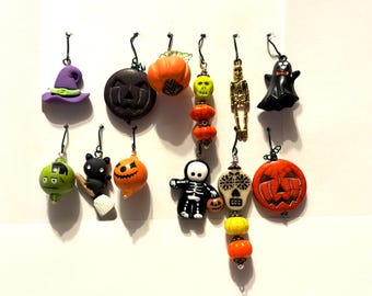 May include: A collection of eleven Halloween-themed charms featuring a purple witch hat, black jack-o'-lantern, orange pumpkin, green skull, gold skeleton, black ghost, green monster head, black cat on a broom, orange pumpkin, skeleton, and skull.