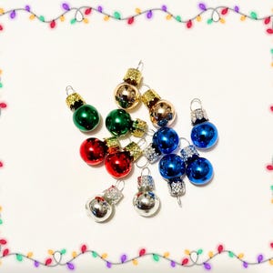 Puede incluir: Una colección de pequeños adornos navideños esféricos en varios colores brillantes, incluyendo rojo, verde, azul, dorado y plateado. Cada adorno cuenta con una tapa metálica con un pequeño lazo para colgar, dispuestos sobre un fondo blanco.