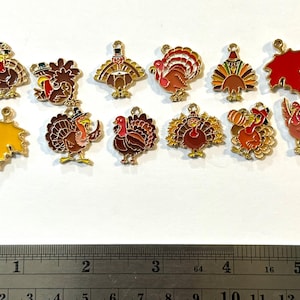 May include: A collection of Thanksgiving-themed charms. The charms feature colorful turkey designs with gold-tone accents, along with two leaf charms. The charms are arranged on a white surface, with a ruler at the bottom.