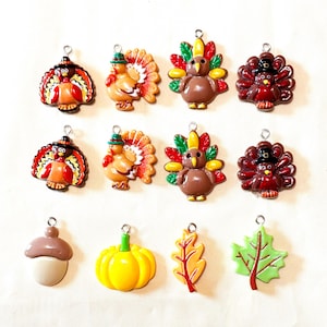 May include: A collection of twelve small charms featuring Thanksgiving themes, including turkeys with colorful feathers and hats, a brown acorn, a bright yellow pumpkin, an orange oak leaf, and a light green maple leaf, each with a metal loop.