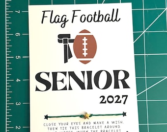 Puede incluir: Una tarjeta rectangular blanca con el texto Flag Football SENIOR 2027, un gráfico de balón de fútbol americano y una pulsera de hilo verde con un pequeño dije de estrella dorada. La tarjeta incluye instrucciones impresas sobre deseos.