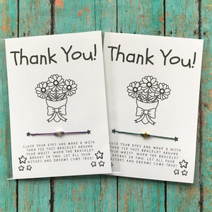 May include: Two white cards with "Thank You!" in black text, each featuring a flower bouquet illustration and a wish bracelet. One bracelet is purple with a heart charm, the other is green with a star charm.