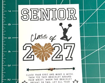 Puede incluir: Una tarjeta rectangular blanca con el texto SENIOR CLASS OF 2027, un corazón de pompones dorados y la silueta negra de una animadora. Se incluye una pulsera de hilo negro con un pequeño dije de corazón y las instrucciones impresas debajo.
