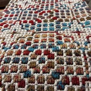 May include: Close-up of a woven textile with a grid pattern. The design incorporates squares of red, blue, and brown hues, set against a white background. The texture appears soft and inviting, suggesting a decorative item.