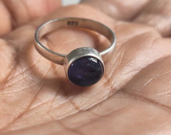 May include: A silver ring featuring a dark purple, circular gemstone. The ring has a plain band and a bezel setting. The ring is displayed on a person's palm. The ring is marked with the number 925.