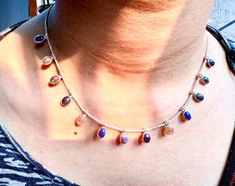 May include: A silver necklace with small, oval-shaped gemstones in various colors, including blue, purple, and iridescent hues. The gemstones are evenly spaced along the delicate chain, creating a sparkling and elegant accessory.