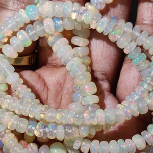 Peut inclure: Un collier de perles d'opale translucides blanc cassé, de forme ronde et aplatie. Chaque perle présente un jeu de couleurs vibrant, avec des éclats iridescents de vert, bleu, orange et rose visibles sur toute la surface de la pierre.