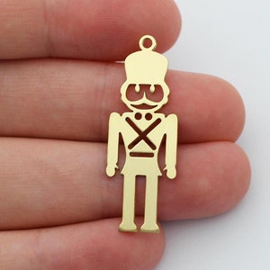 May include: Two gold-coloured metal nutcracker charms. Each charm features a detailed silhouette of a nutcracker soldier with a hat, moustache, and crossed straps.