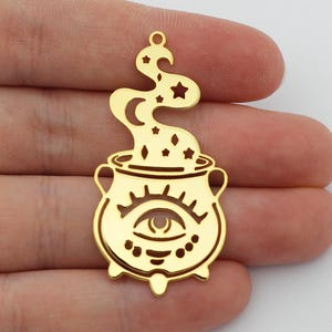 Puede incluir: Encanto de metal dorado en forma de caldero con un solo ojo y estrellas en el vapor que sale de la parte superior.