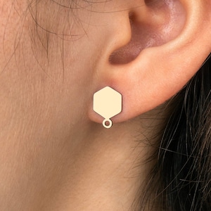 May include: Gold hexagon stud earrings with a small dangling circle. The earrings are worn in a person's ear.