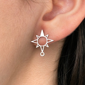 Pode incluir: Um par de brincos prateados. Cada brinco apresenta um design estilizado de estrela solar com oito pontas e um pequeno elemento pendurado. Os brincos são mostrados em uma orelha.