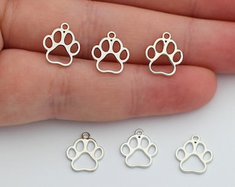 May include: Six small silver-toned metal charms shaped like animal paw prints. Each charm features a hollow centre and a small circular loop at the top for attachment. The charms are arranged with three resting on a palm and three below.