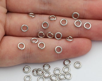 Könnte beinhalten: Eine Ansammlung kleiner, kreisförmiger silberfarbener Metall-Biegeringe. Diese Schmuckzubehörteile sind dünne, offene Schlaufen, die zum Verbinden von Komponenten in Bastelprojekten dienen. Die Ringe sind verstreut und liegen in einer Handfläche.