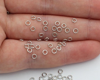 Könnte beinhalten: Eine Sammlung kleiner, kreisförmiger silberfarbener Metall-Biegeringe. Diese Schmuckzubehörteile sind dünne, offene Schlaufen, die zum Verbinden von Komponenten in Bastelprojekten verwendet werden. Die Ringe liegen verstreut in einer offenen Handfläche.