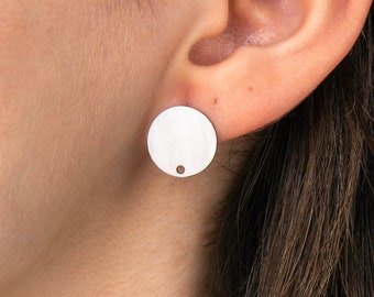 May include: A circular silver-toned stud earring featuring a smooth, flat surface with a small, distinct hole positioned near the bottom edge. The earring is secured in the earlobe, displaying a metallic finish and a simple, geometric design.