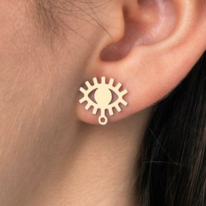 Puede incluir: Pendientes dorados en forma de ojo con un pequeño colgante. Los pendientes se llevan en la oreja de una persona.