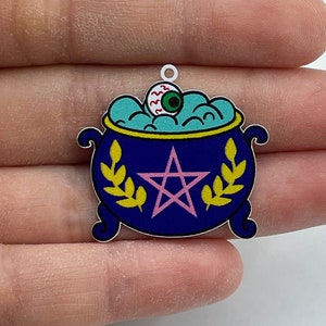 Puede incluir: Un amuleto de caldero azul con un pentagrama, hojas doradas y un ojo flotando. El amuleto está hecho de metal y tiene un acabado plateado.