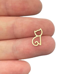 May include: A small, gold-colored metal charm in the shape of a stylized cat silhouette. The design features a simple outline of a cat head with pointed ears and a curved tail, creating a minimalist, open-frame cat figure made of thin metal wire.