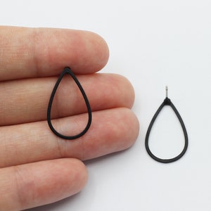 Op de afbeelding: Twee zwarte, druppelvormige oorbelcomponenten. Eén wordt in een hand gehouden, de andere ligt op een wit oppervlak. De oorbellen hebben een eenvoudig, minimalistisch ontwerp, met een dunne omtrek en een pin voor bevestiging. De oorbellen zijn waarschijnlijk voor het maken van sieraden.