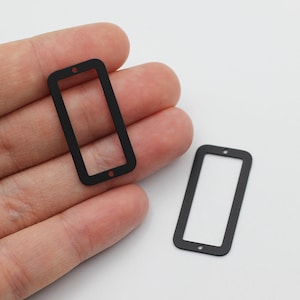 May include: Two matte black rectangular metal frames with rounded corners and a small hole at the top and bottom centre of each piece. These flat, thin components are designed for crafting or jewellery making, featuring a simple open centre.