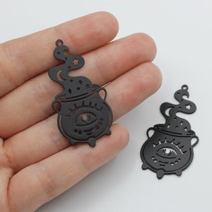 Puede incluir: Dos encantos de metal negro con forma de caldero con un solo ojo y estrellas en el vapor.