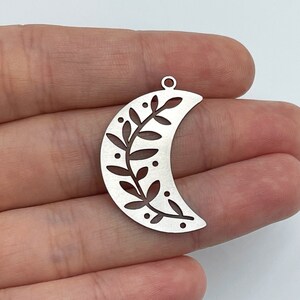 May include: A silver-toned crescent moon pendant featuring a cut-out leaf and vine pattern with small circular accents. The metal charm has a smooth, matte finish and includes a small circular loop at the top for attachment to a chain or cord.