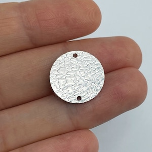 以下が含まれることがあります： テクスチャ加工された表面と、ジュエリーやその他のクラフトに取り付けるための2つの穴が付いた、銀色の丸いチャームです。