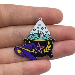Puede incluir: Un amuleto de metal blanco con un caldero negro lleno de líquido azul y un ojo rosa. Un sombrero de bruja morado con medias lunas amarillas se encuentra en el lado del caldero.