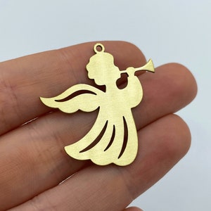 May include: A flat, gold-colored metal charm in the shape of an angel playing a trumpet. The angel figure features a circular head, outstretched wings, and a flowing gown with cut-out details. A small circular loop is located at the top for hanging.