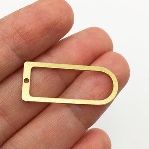May include: A gold-colored metal pendant with a rectangular shape and a rounded end. The pendant has a small hole on one side and is held in a hand against a white background. This jewelry component is suitable for crafting.