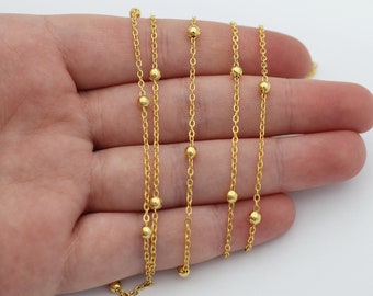 May include: A gold-toned chain necklace featuring small, evenly spaced spherical beads integrated along the length of the delicate metal links. The chain is draped in multiple sections, showcasing the consistent metallic finish and bead placement.