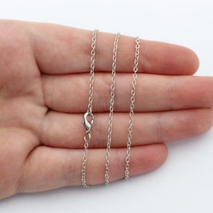 May include: A silver chain necklace with a lobster clasp closure. The chain is shown on a hand, with the chain laying flat.