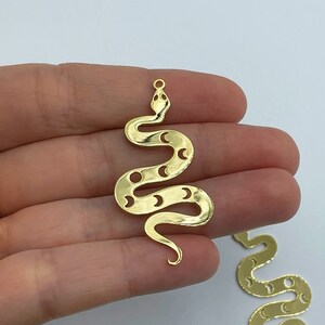 Pode incluir: Um pingente de cobra em metal dourado com uma série de formas de lua crescente recortadas ao longo do corpo. A cobra é representada em uma postura sinuosa em forma de S com um pequeno laço circular no topo para fixação, com superfície lisa.