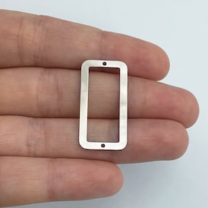 Può includere: Un connettore rettangolare in metallo color argento con angoli arrotondati e un centro cavo. Il pezzo presenta due piccoli fori circolari posizionati sui bordi superiore e inferiore, progettati per il fissaggio in progetti di gioielleria.