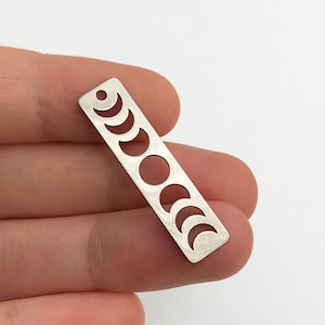 May include: Silver charm with a cutout design of the phases of the moon. The charm is rectangular and has a hole at the top for attaching to a bracelet or necklace.