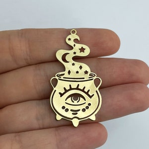 Puede incluir: Encanto de metal dorado con un diseño de caldero. El caldero tiene un solo ojo con una luna creciente en su interior y estrellas alrededor. Del caldero se eleva humo con estrellas y una luna creciente en el humo.